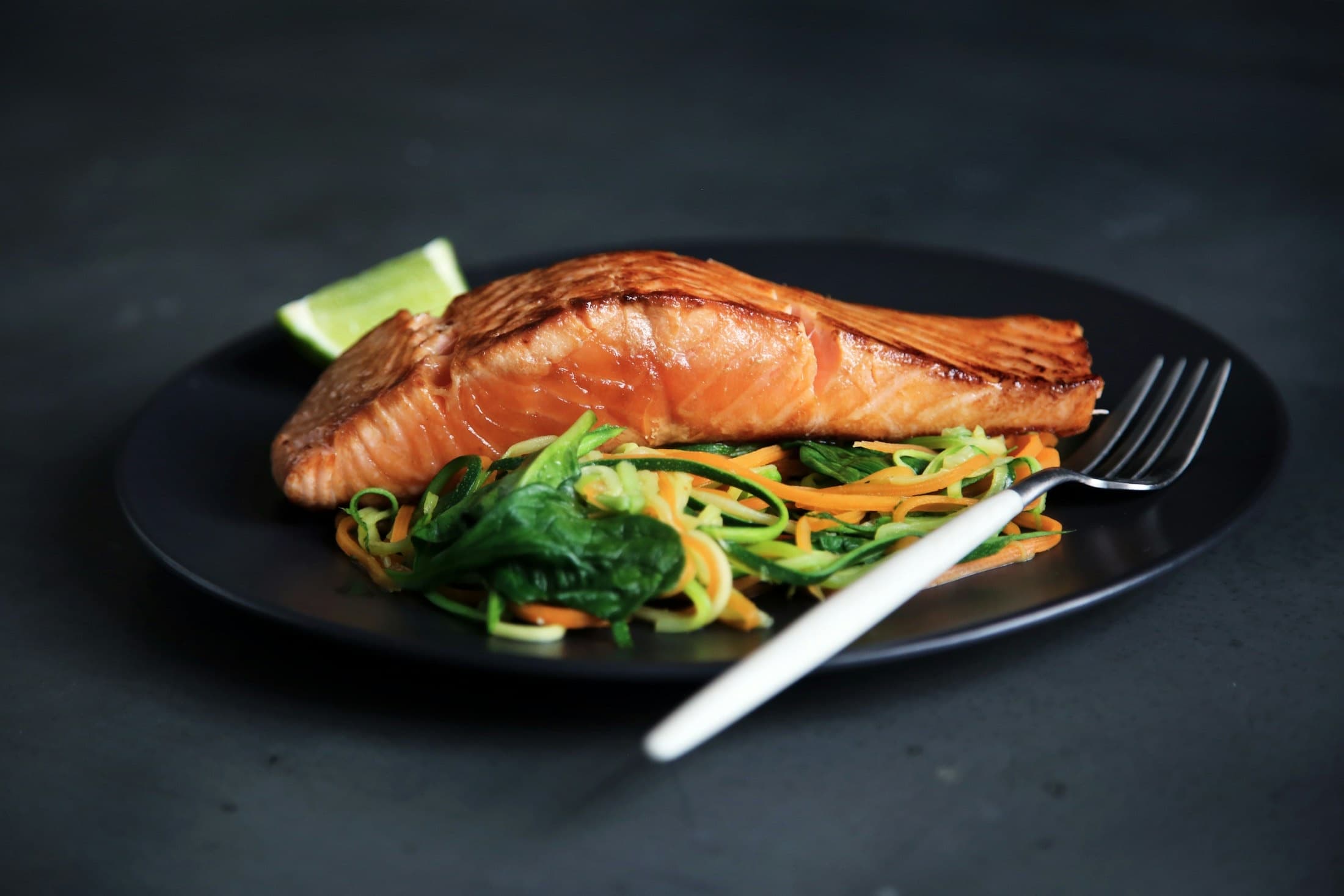 Seared salmon fillet on a dark plate with spiralized zucchini and carrot, lit by dramatic side light