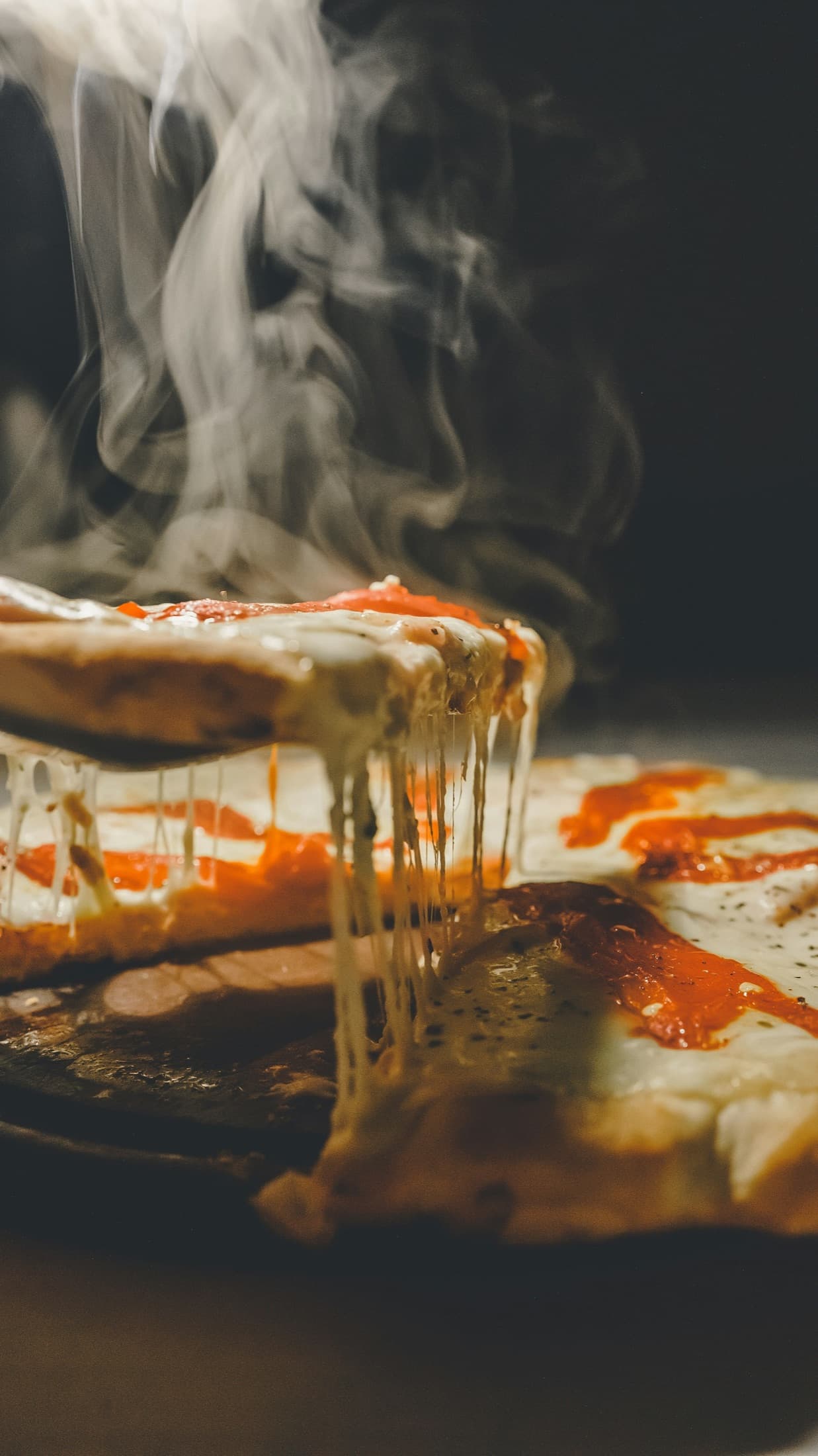 A hot slice of pizza being lifted from the pie, steam rising, on a dark surface